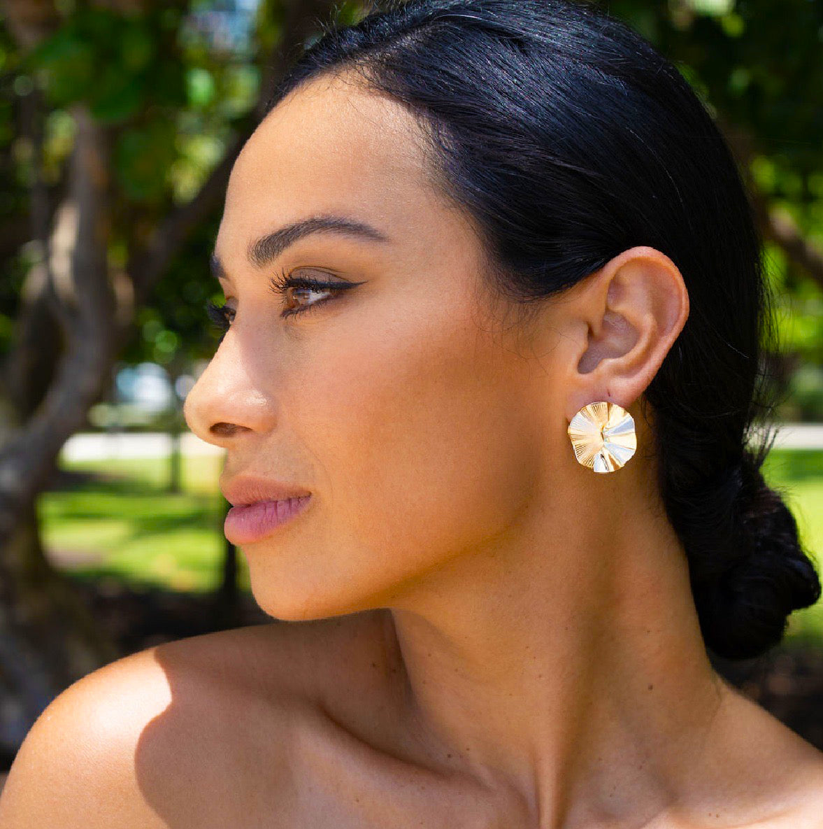 A woman with dark hair in an updo wears large, textured gold earrings while posing outdoors in natural sunlight.