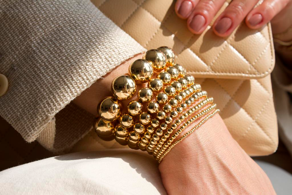 A woman wears a stylish stack of shiny gold ball bead bracelets on her wrist while holding a beige quilted handbag.