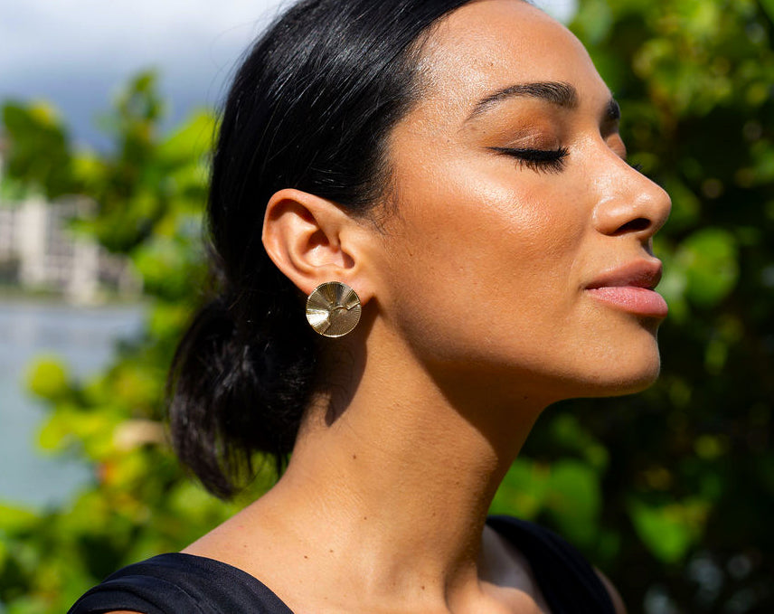 A serene woman wearing textured gold statement earrings turns her head outdoors against a lush green background.
