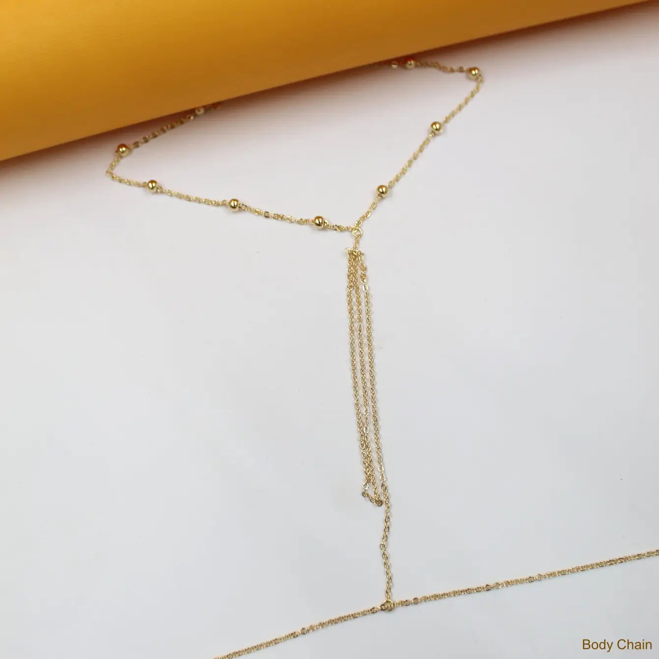 A delicate gold-beaded body chain rests on a clean white surface against an orange backdrop.