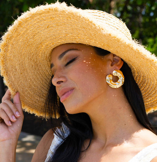 A glamorous woman wearing a large, textured gold hoop earring poses outdoors in a sunlit straw hat.