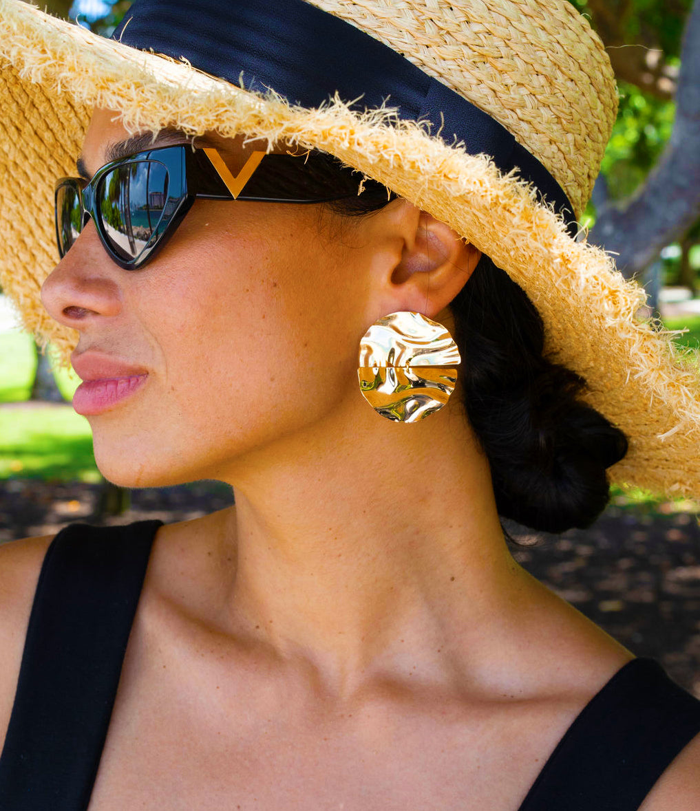 A stylish woman wearing a straw sun hat and chic sunglasses showcases textured gold statement earrings outdoors.