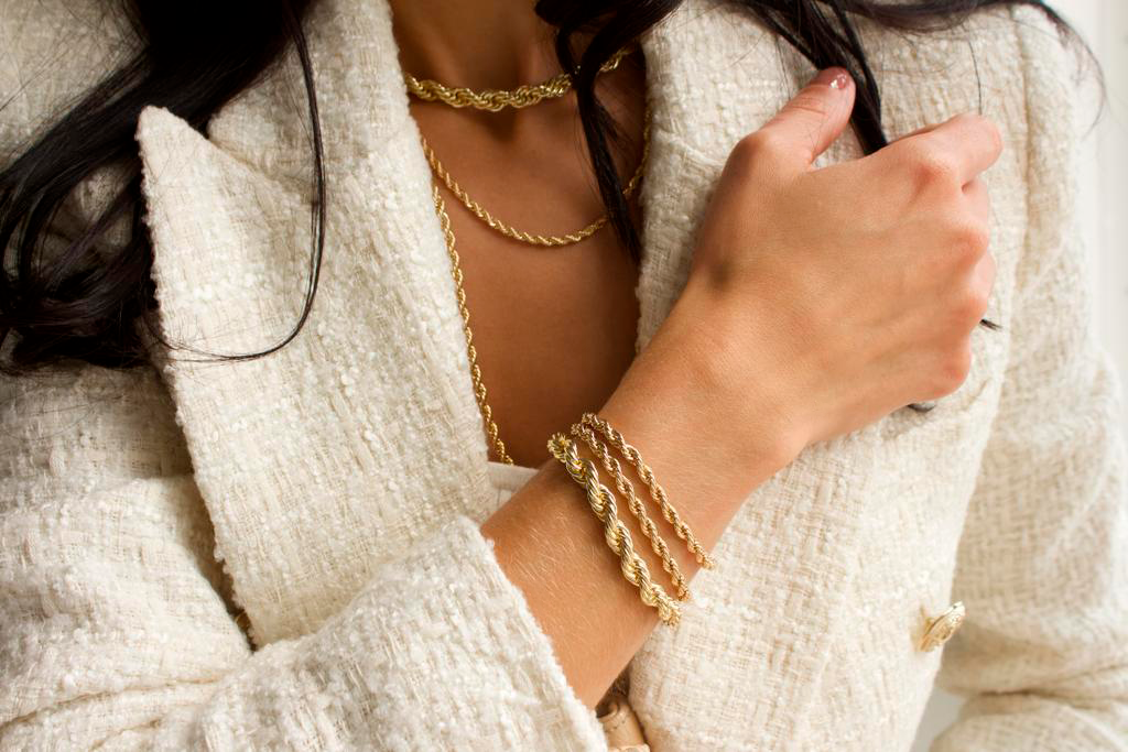 A stylish woman wearing a textured tweed blazer models layered gold rope necklaces and matching gold rope bracelets.