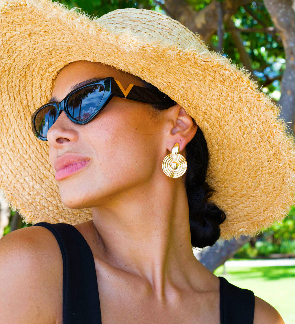 A stylish woman wearing a large straw sun hat, black sunglasses, and textured gold disc earrings poses outdoors on a sunny day.