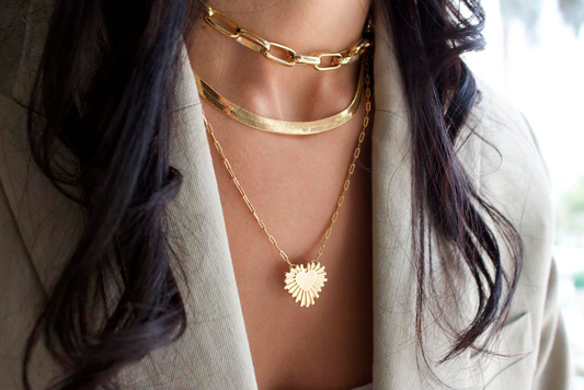 A woman wears layered gold necklaces, including a chunky chain choker, a herringbone chain, and a pendant necklace with a sunburst heart charm.