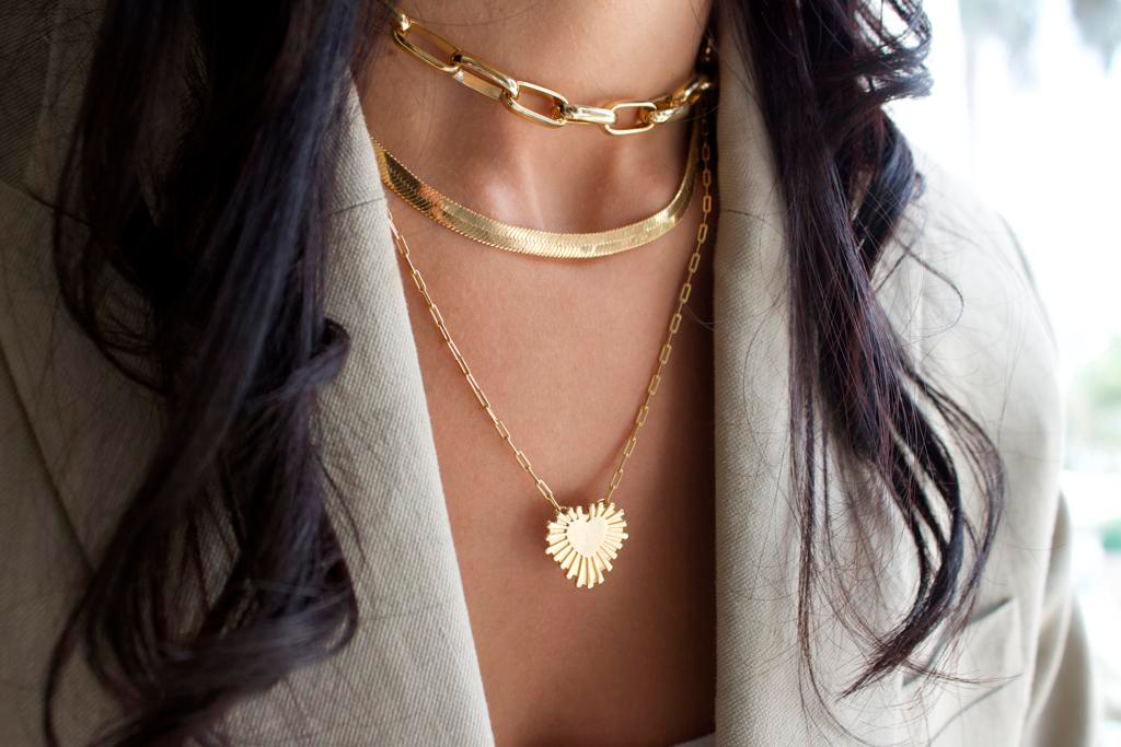 A stylish woman wears a layered gold chain necklace set featuring a chunky choker, a flat herringbone chain, and a radiant heart pendant while wearing a beige blazer.