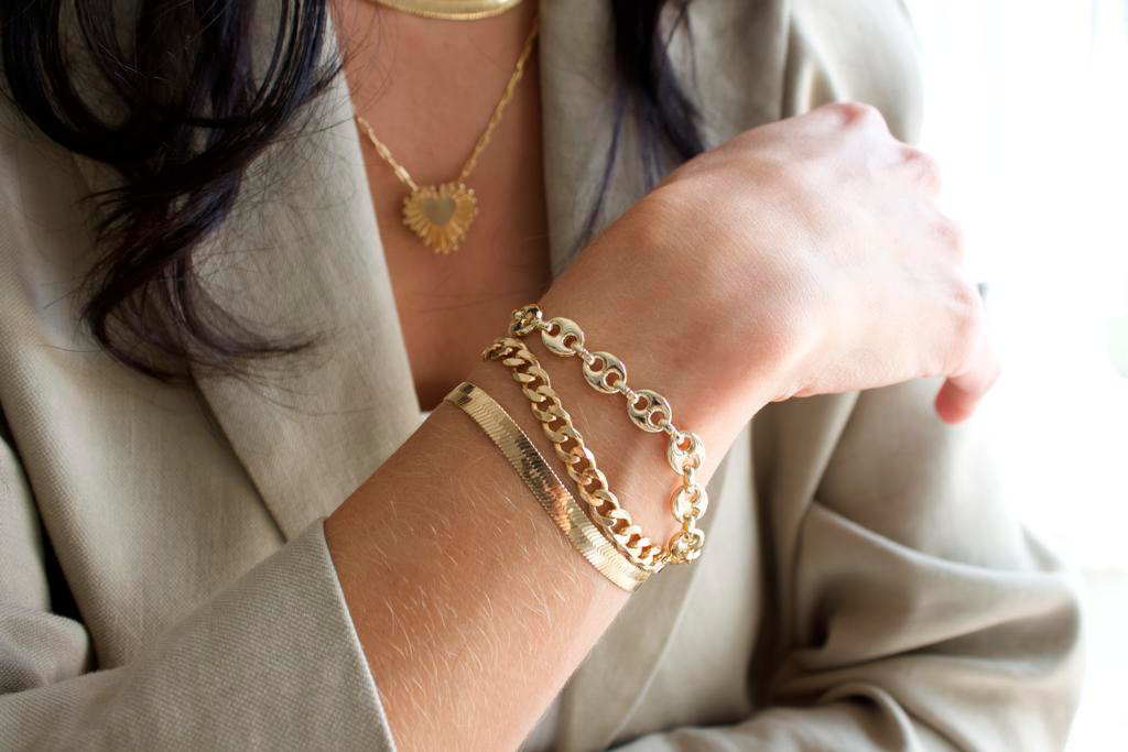 A woman wearing a beige blazer models a stacked gold bracelet set featuring a herringbone chain, curb chain, and mariner link bracelet.