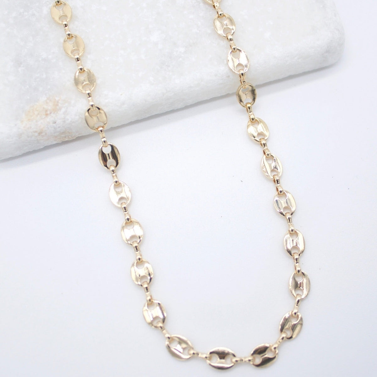 A shiny gold mariner chain necklace drapes across a white marble surface against a clean background.