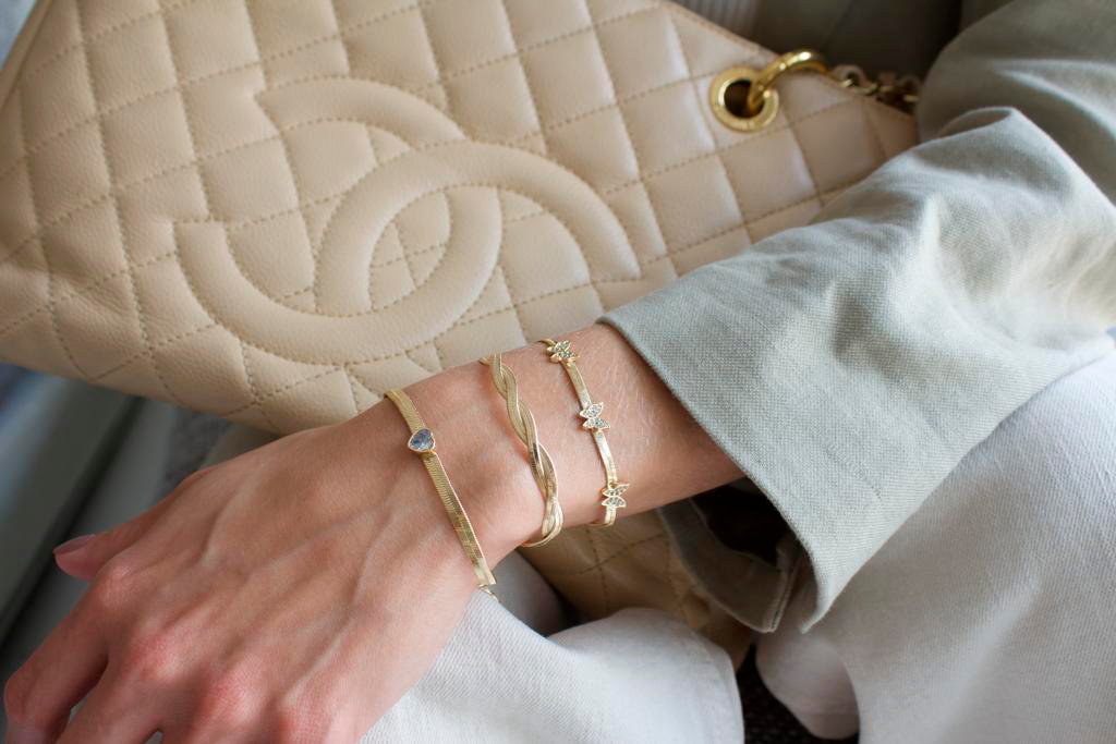 A woman wearing three stacked gold bracelets showcases her wrist against a quilted beige handbag.
