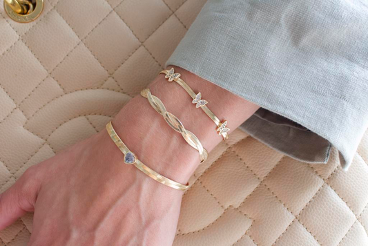 A person wears three stacked gold herringbone bracelets featuring a butterfly charm, a twisted design, and a heart accent against a quilted handbag.