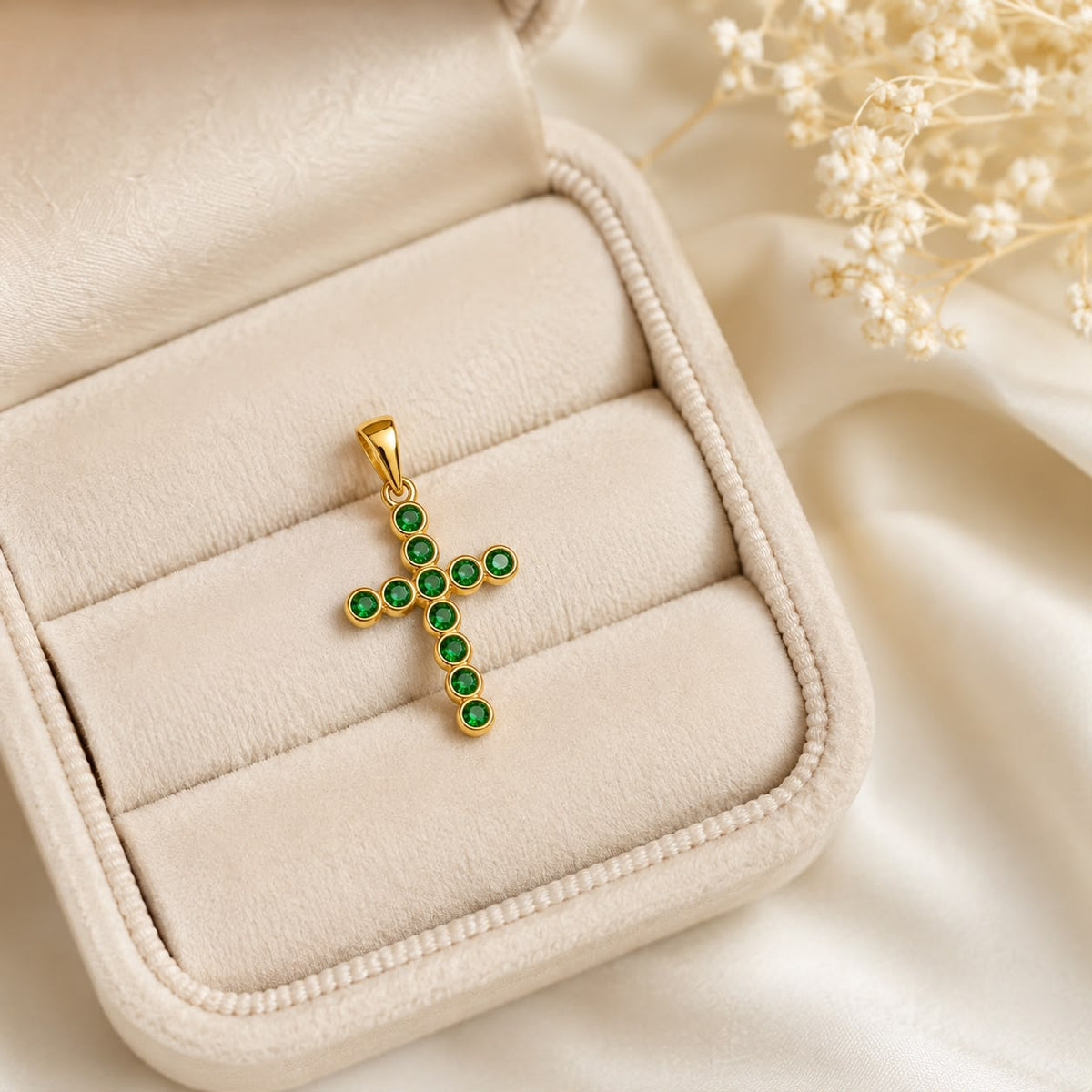 A gold cross pendant encrusted with vibrant green gemstones rests neatly inside an open cream-colored velvet jewelry box.