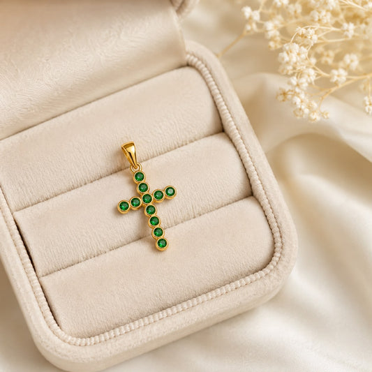 A gold cross pendant encrusted with vibrant green gemstones rests neatly inside an open cream-colored velvet jewelry box.