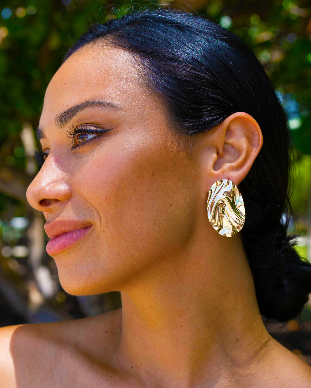 A woman with dark hair tied back showcases a pair of large, abstract gold earrings outdoors.