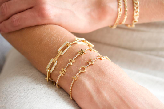 A person wears a layered stack of gold chain link and diamond-accented initial bracelets, spelling out "MAM" and "LOVE" on their wrist.
