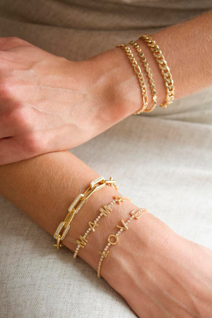 A person models a stylish layered stack of gold chain bracelets and pavé-set letter charm bracelets on each wrist.