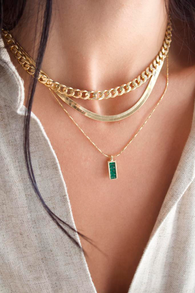 A woman wears a layered gold jewelry set featuring a chunky chain choker, a sleek herringbone chain, and a delicate pendant necklace with a green rectangular stone.