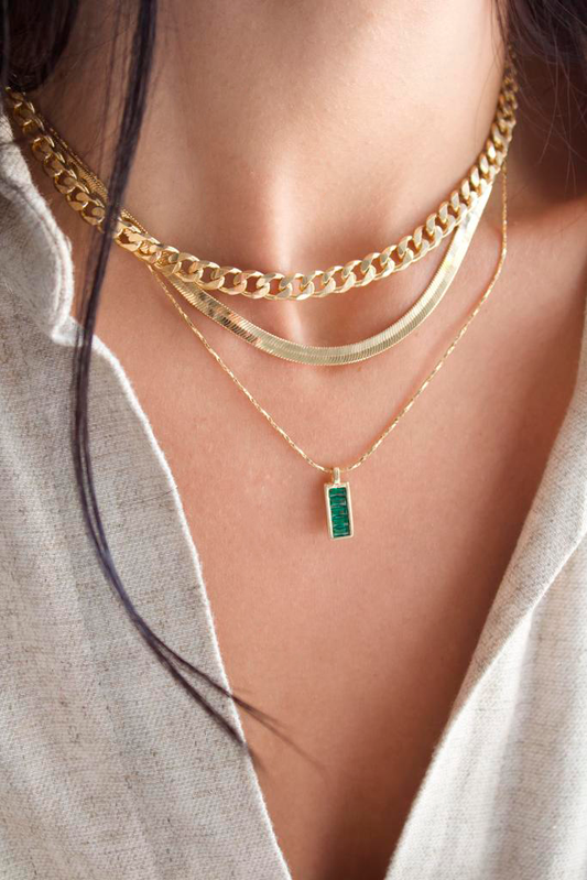 A woman wears a layered gold jewelry set featuring a chunky chain choker, a sleek herringbone chain, and a delicate pendant necklace with a green rectangular stone.