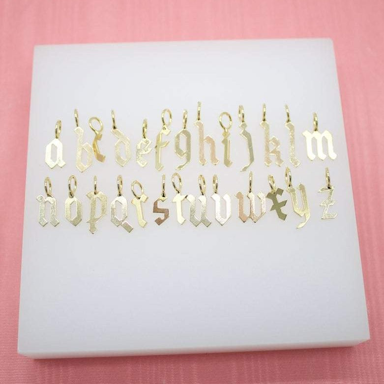 A collection of gold Old English letter charms is neatly arranged in alphabetical order on a white display block.