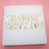 A collection of gold Old English letter charms is neatly arranged in alphabetical order on a white display block.