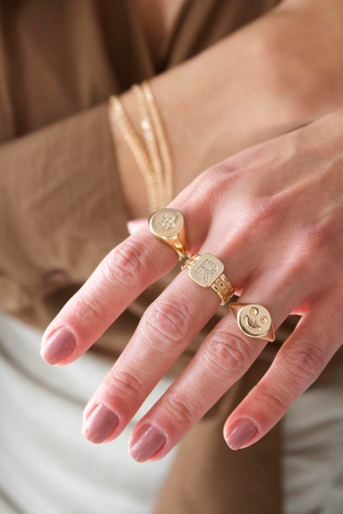 A person wearing layered gold jewelry displays three gold rings on their fingers, featuring a star-shaped signet, an initial R signet, and a smiley face signet.