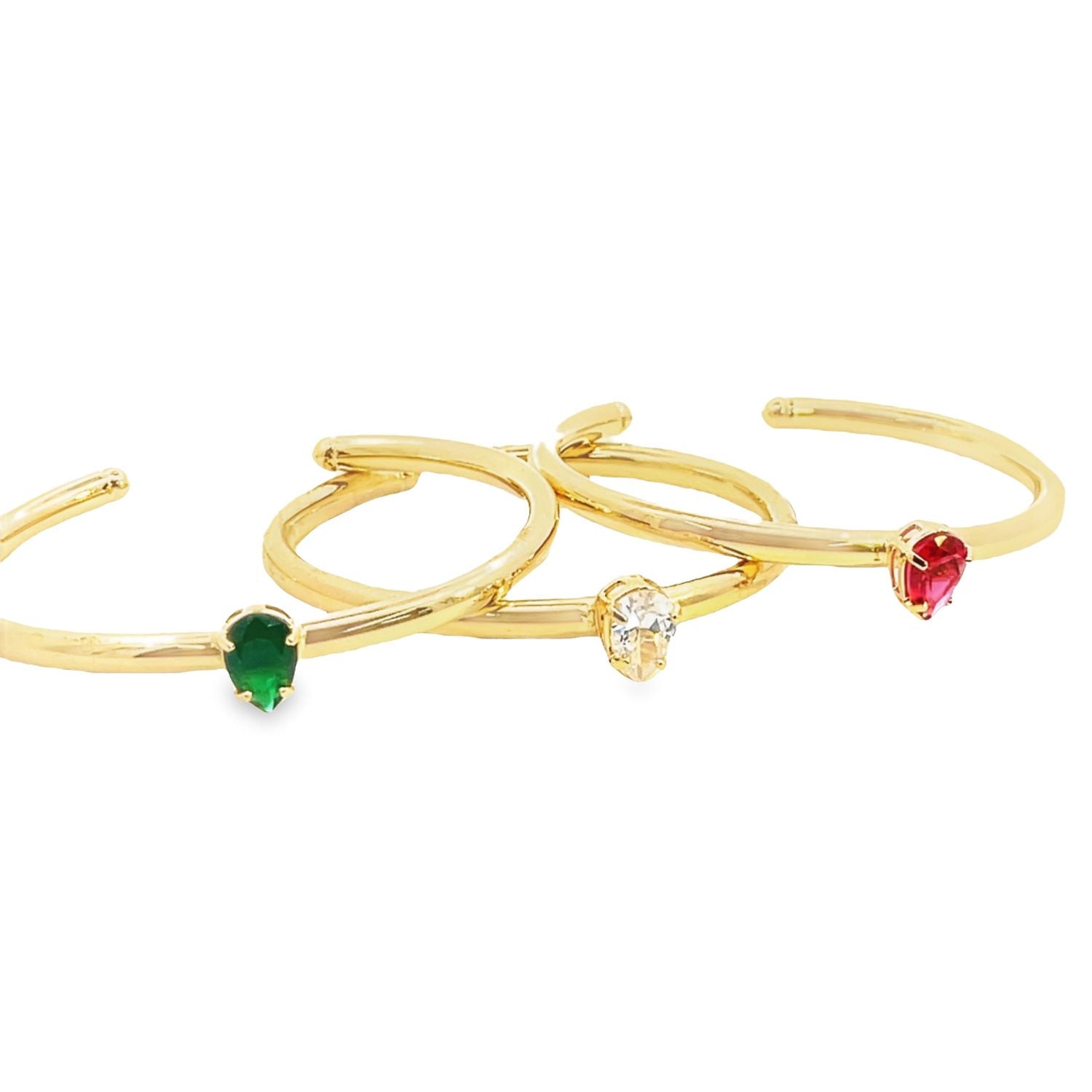 Three gold cuff rings with pear-cut gemstones in green, white, and red rest neatly against a white background.