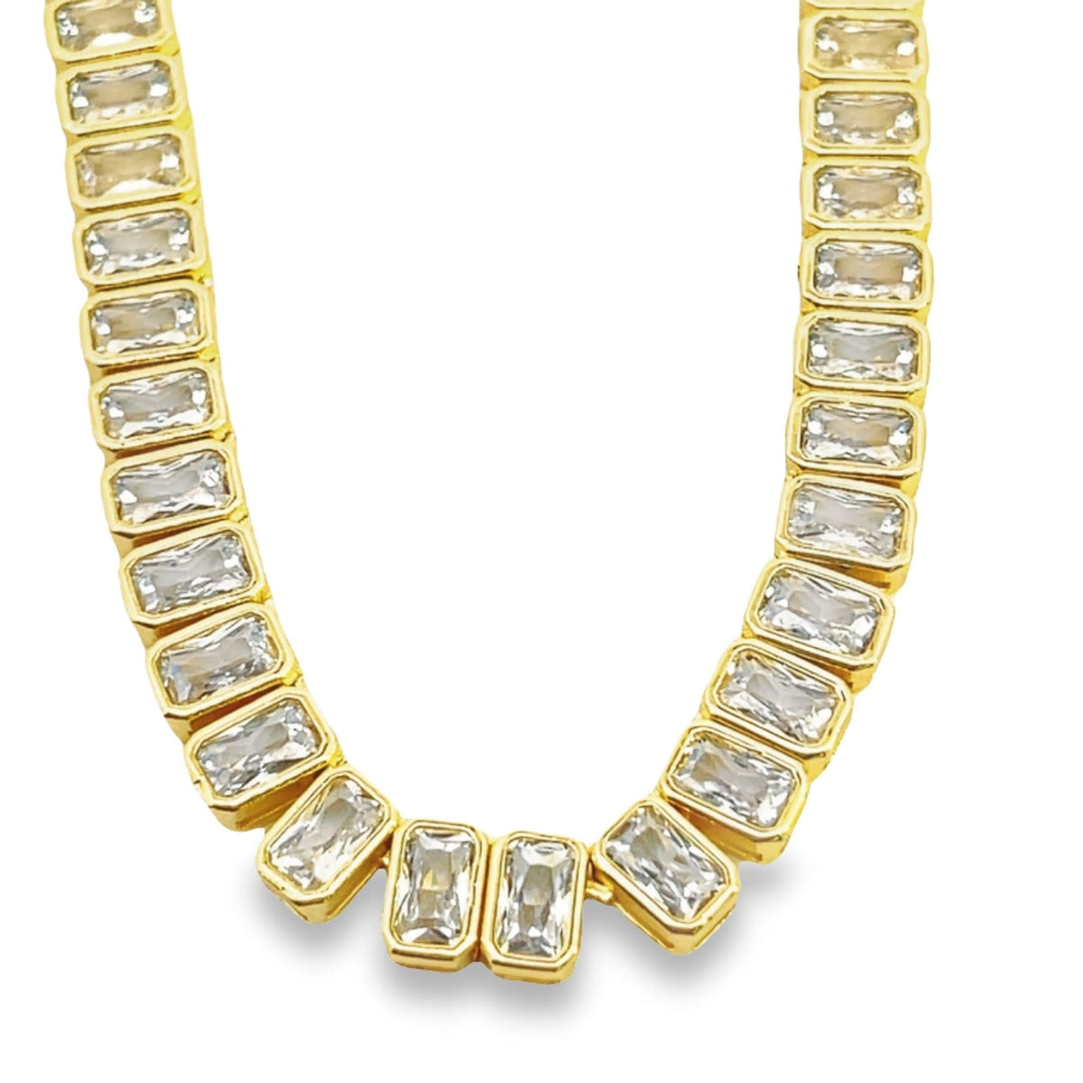 A gold tennis necklace features a continuous strand of bezel-set rectangular crystal stones against a white background.