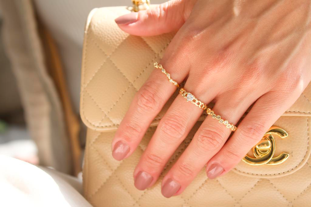 A woman with nude-polished nails wears elegant gold rings with diamonds while holding a beige quilted Chanel handbag.
