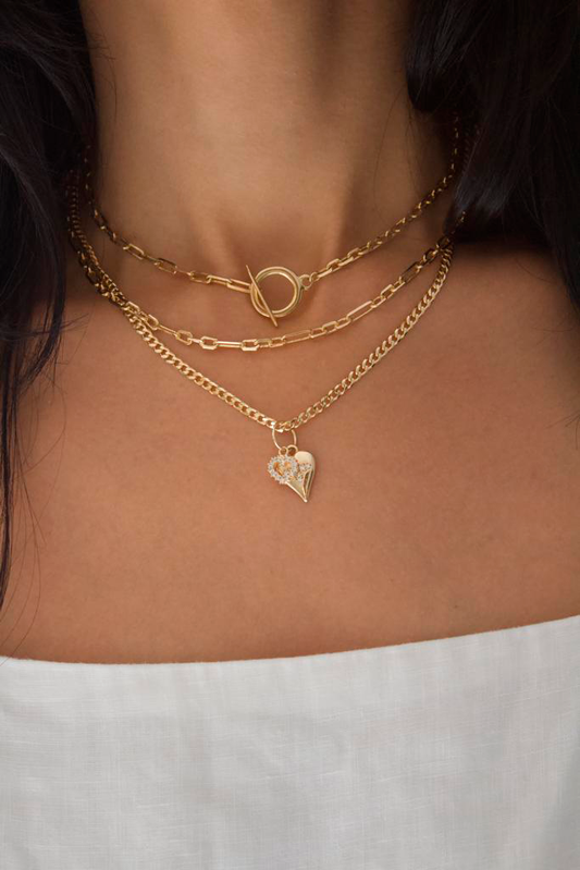 A woman wears a layered gold chain necklace set featuring a toggle clasp and a heart-shaped pendant.