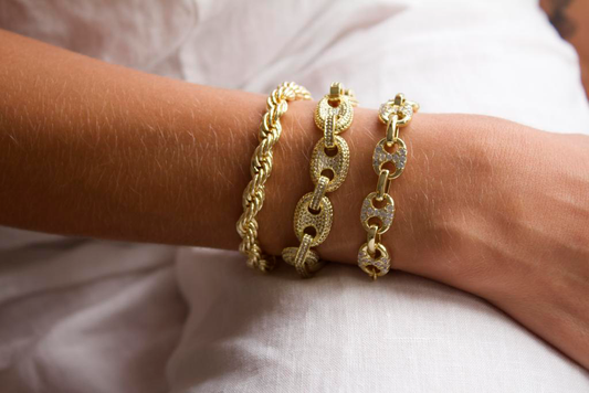 A woman models a stack of three gold bracelets, featuring a thick rope chain, a pave mariner link chain, and a delicate diamond-encrusted chain on her wrist.