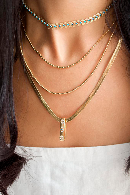 A woman wears four layered gold necklaces, including a choker with blue evil eye details and a herringbone chain with a pendant.