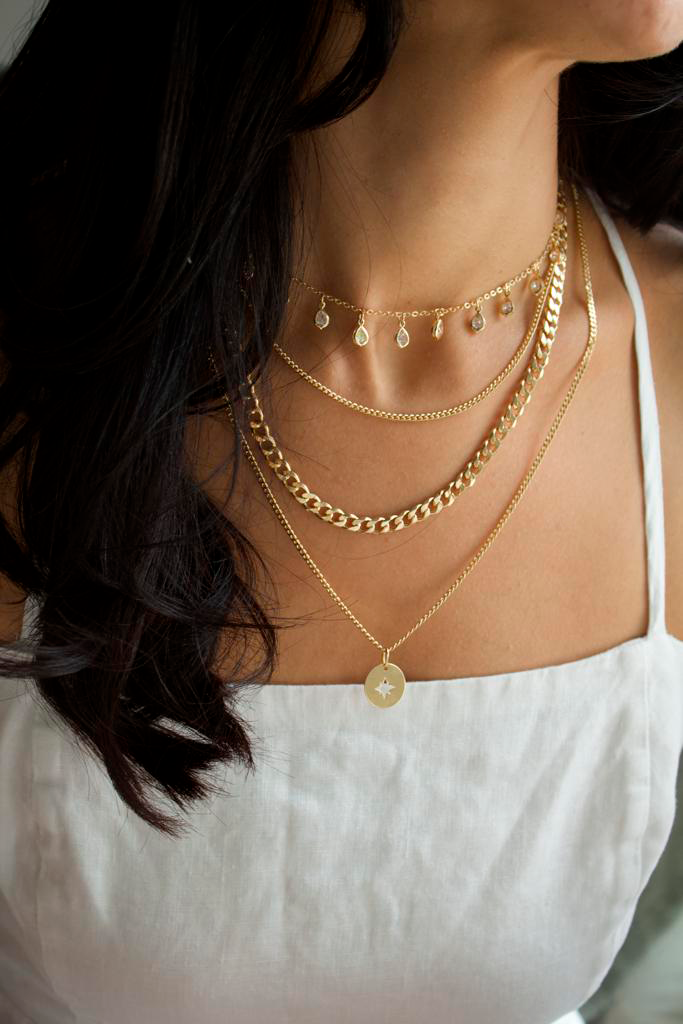 A woman wearing layered gold necklaces, including a pendant choker and a coin pendant necklace, stands in a white dress.