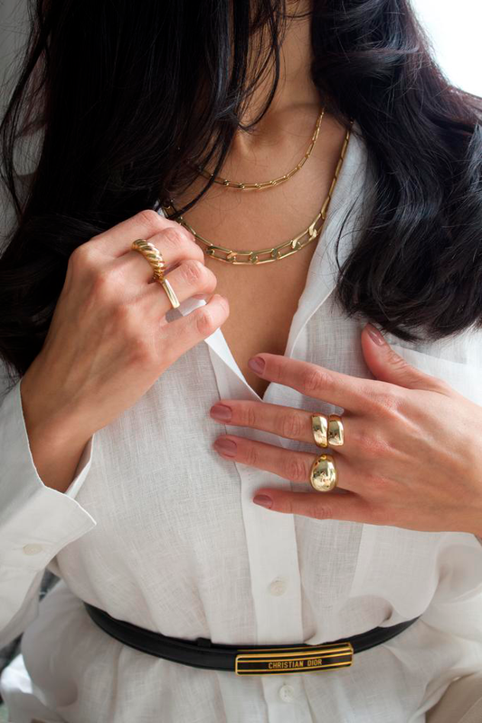 A stylish brunette woman wearing a white button-down shirt and layered gold chain necklaces showcases chunky gold rings on her fingers.