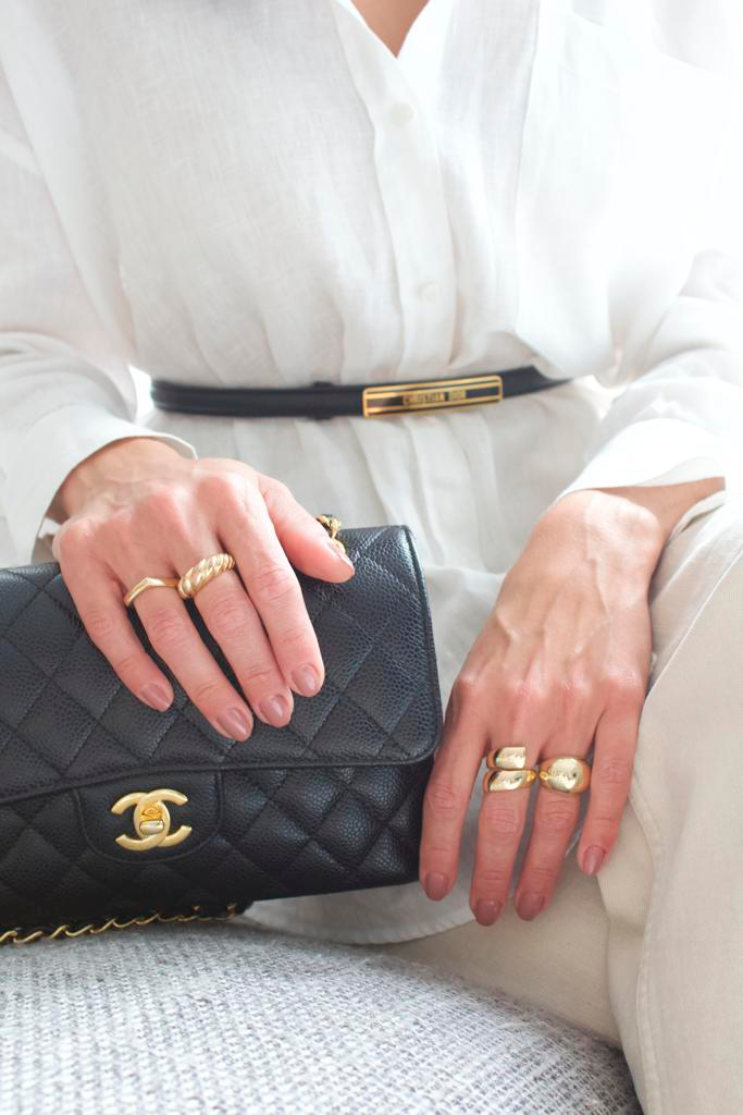 A stylish woman wearing a white linen shirt and gold statement rings rests her hands on a quilted black Chanel handbag.