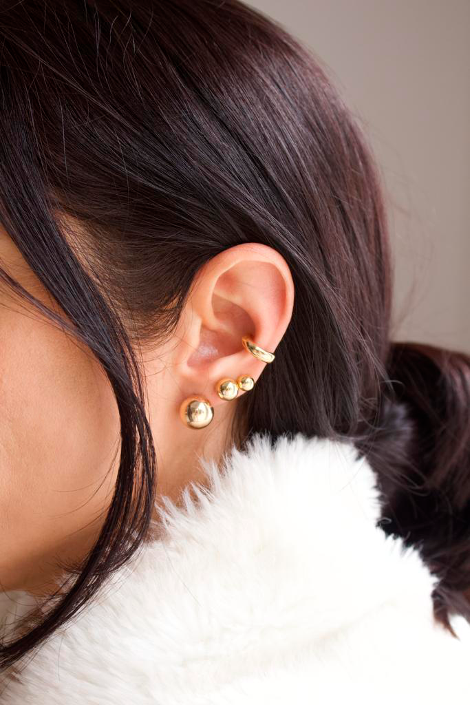 A person models a curated stack of gold earrings, featuring a smooth conch cuff and three graduated ball studs along the earlobe.