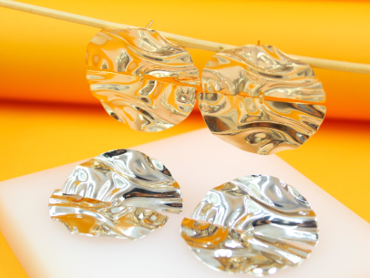 Four shimmering gold-plated and silver-plated textured disc earrings rest on a white surface against a bright yellow background.