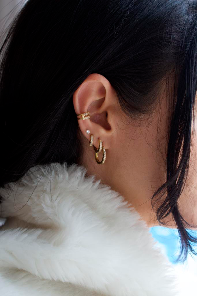 A woman with dark hair displays a curated ear stack featuring gold hoop earrings, a stud, and a crystal-embellished ear cuff.
