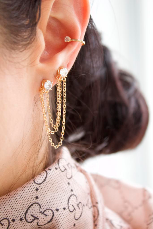 A close-up shot highlights a person's ear adorned with trendy gold chain drop earrings featuring sparkling crystal studs and a matching gold cuff.