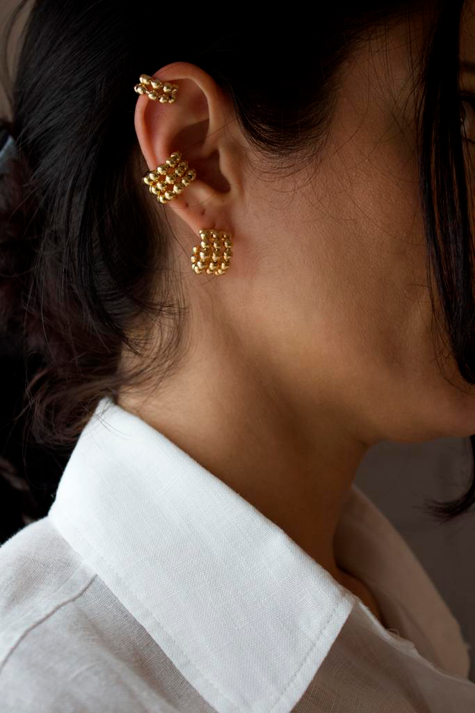 A woman wears a matching set of gold beaded jewelry, including two layered ear cuffs and a small hoop earring.