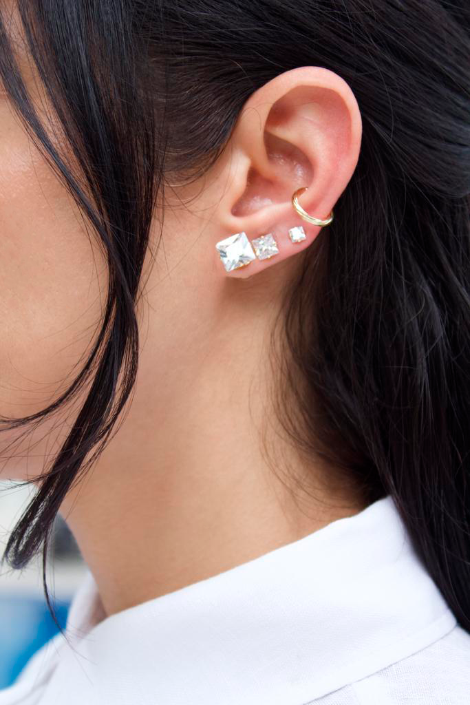 A woman with dark hair showcases an ear stack featuring graduated square crystal stud earrings and a gold conch cuff.