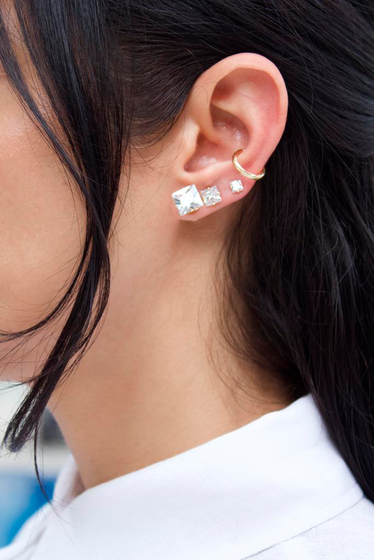 A woman with dark hair showcases an ear stack featuring graduated square crystal stud earrings and a gold conch cuff.