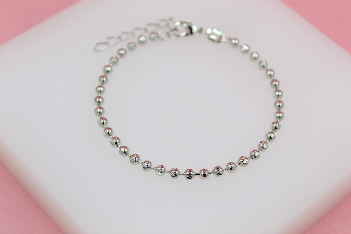 A delicate silver ball chain bracelet rests on a white display surface against a pink background.