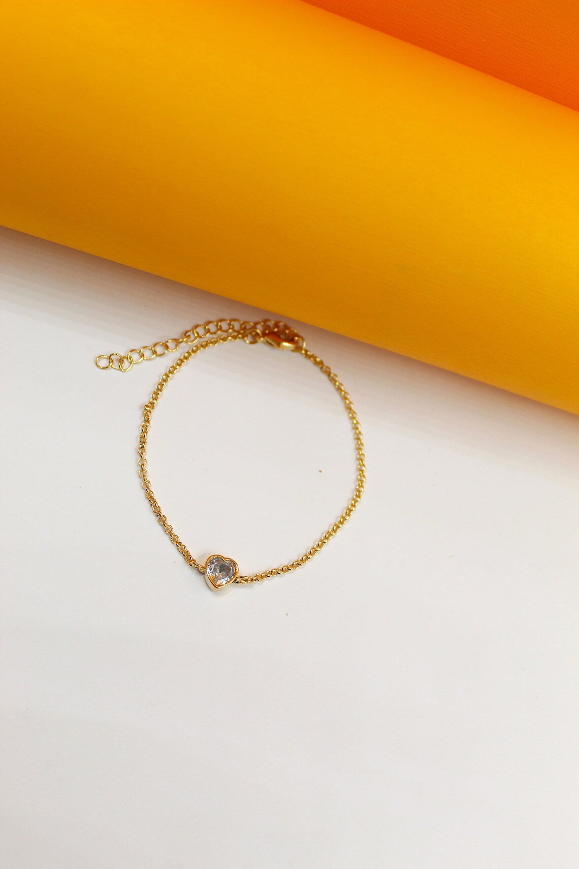 A delicate gold chain bracelet features a sparkling heart-shaped gemstone centerpiece against a vibrant yellow and white background.