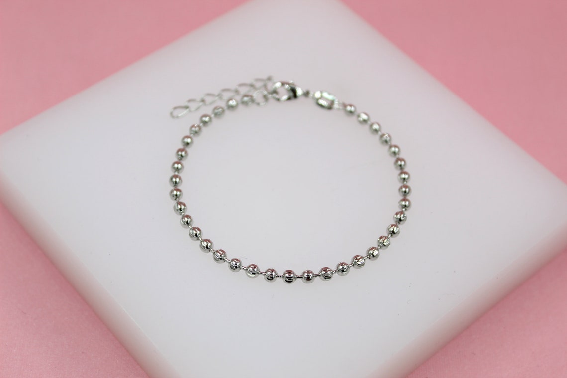 A delicate silver ball chain bracelet rests on a white display stand against a pink background.