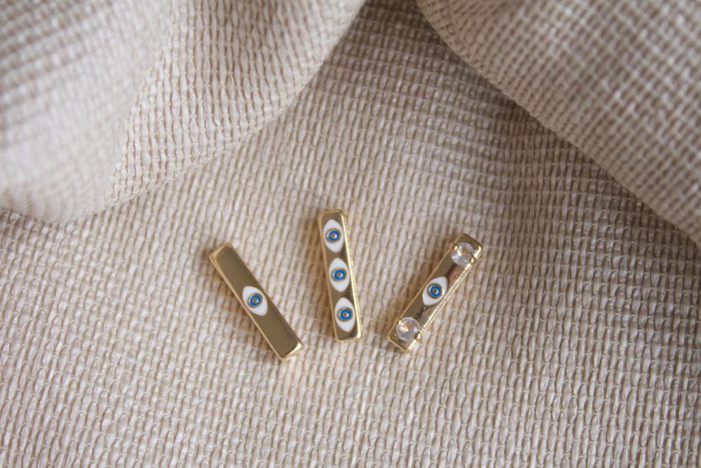 Three gold bar charms featuring blue evil eye symbols and sparkling crystals rest on a textured beige fabric.