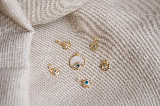 Several gold evil eye pendants rest on a textured beige fabric background.
