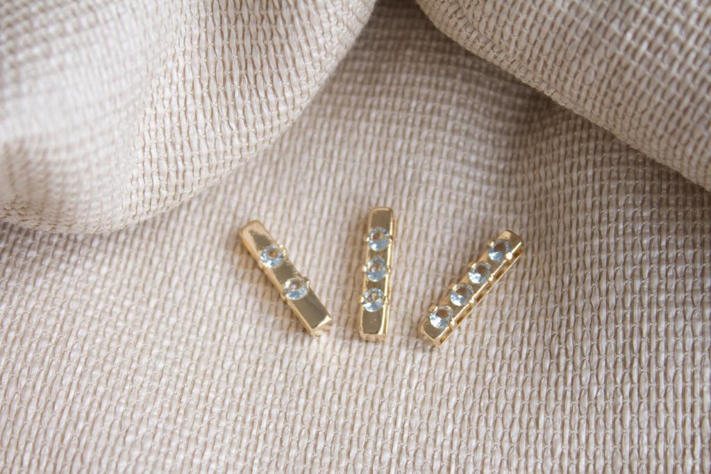Three delicate gold bar earrings set with sparkling light blue gemstones rest on a textured neutral fabric backdrop.