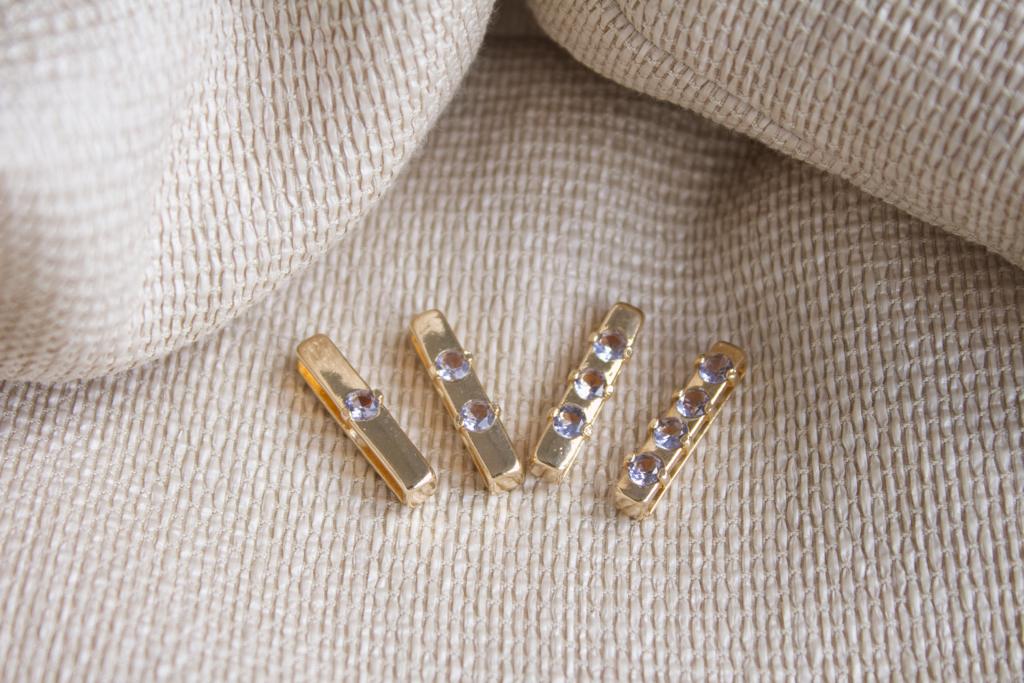 Four gold bar charms, each embedded with sparkling purple gemstones, rest neatly on a textured beige fabric background.