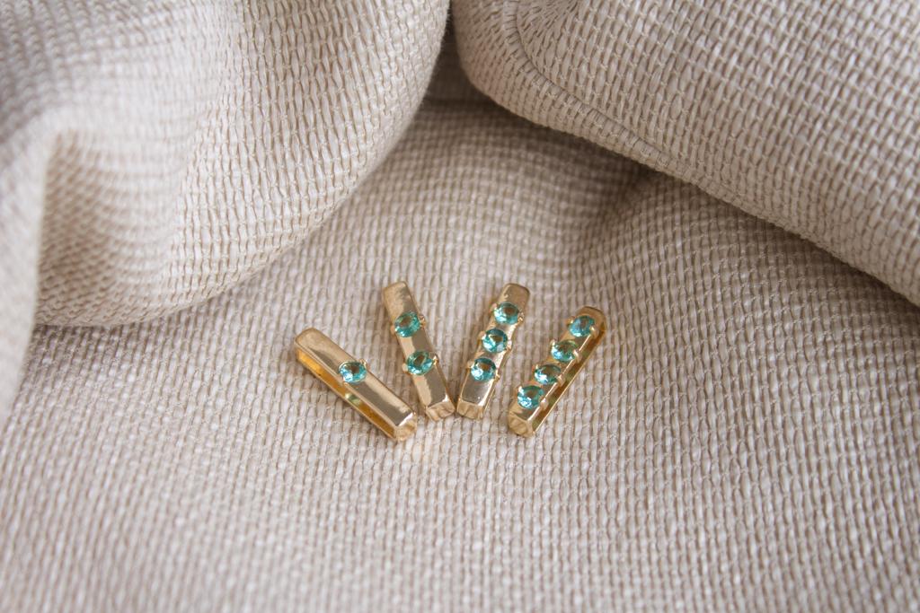 Four gold bar charms encrusted with sparkling turquoise gemstones rest on a textured beige fabric.