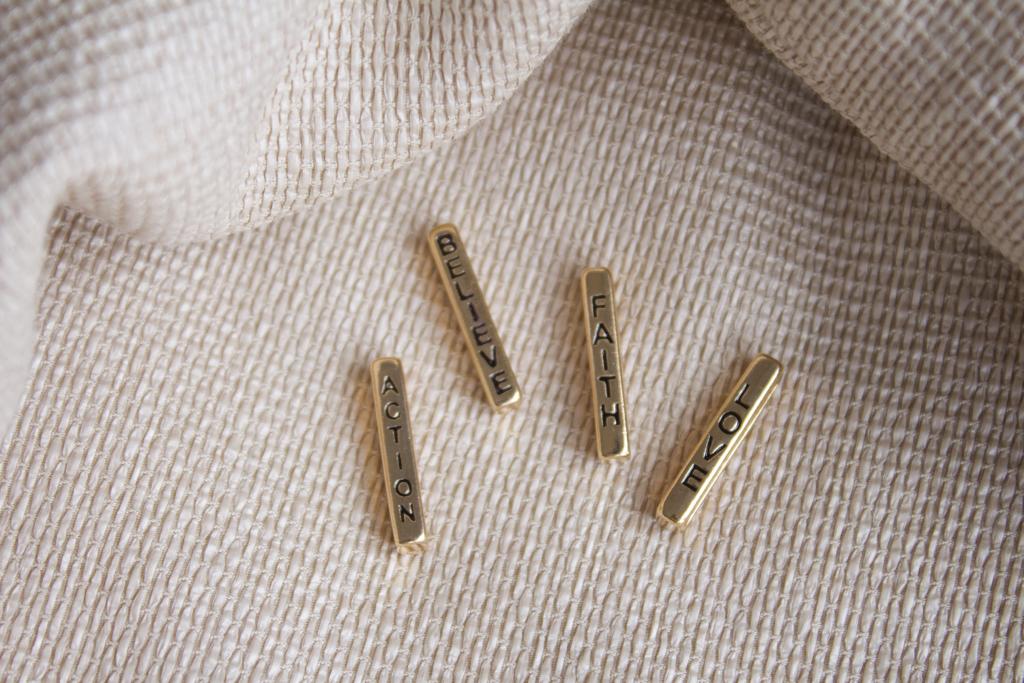 Four gold bar beads engraved with the words "Action," "Believe," "Faith," and "Love" rest on a textured linen background.