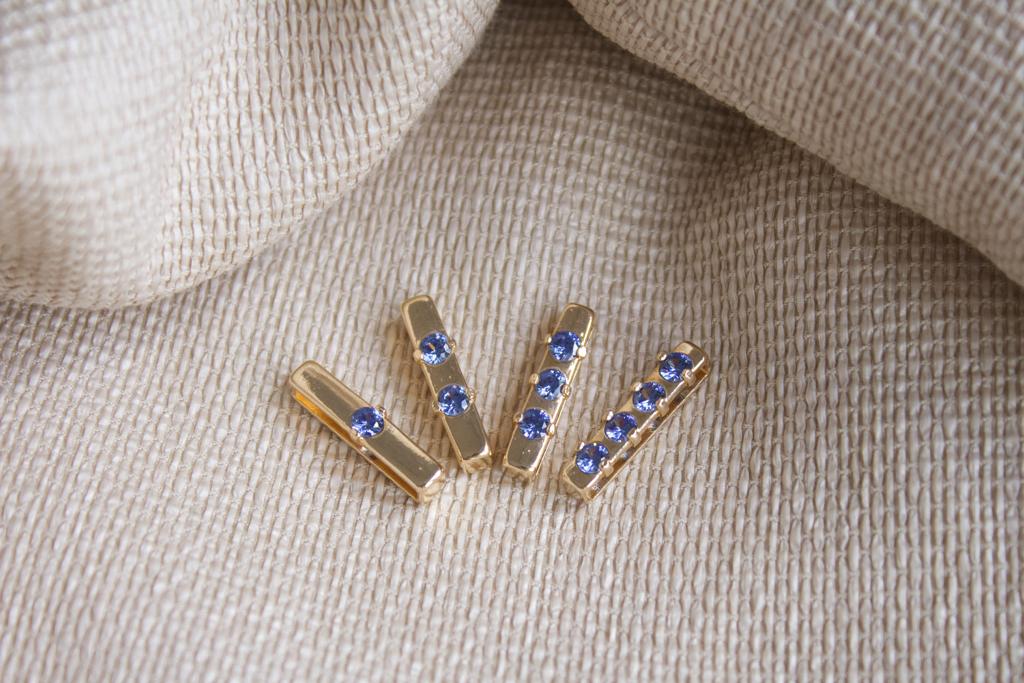 Four gold bar pendants featuring one to four brilliant-cut blue sapphire gemstones rest on a textured beige fabric background.